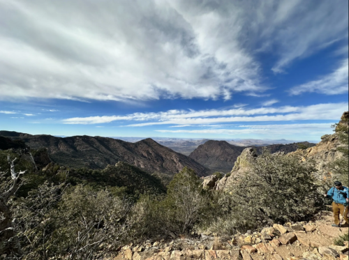 A hiker stands on a rocky trail overlooking a vast mountain landscape under a blue sky with scattered clouds.