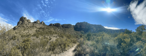 Panoramic view of rugged mountains under a bright blue sky with scattered clouds and sun shining. Lush greenery in foreground.