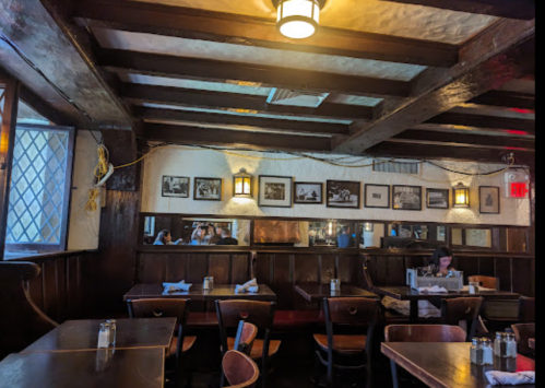 Cozy restaurant interior with wooden beams, tables, and framed photos on the walls; a lone diner is seated.