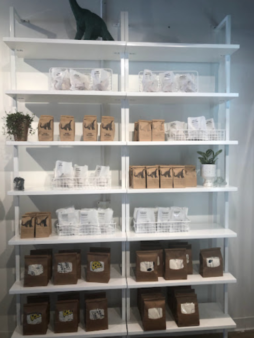 A white shelf displays various bags of products, including brown and white packaging, with a small plant and dinosaur figurine.