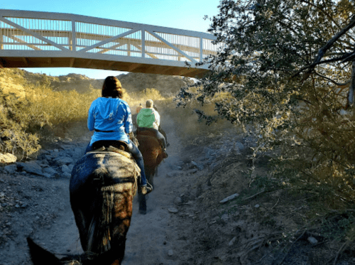 Two riders on horseback travel along a dusty trail beneath a wooden bridge, surrounded by sparse vegetation.