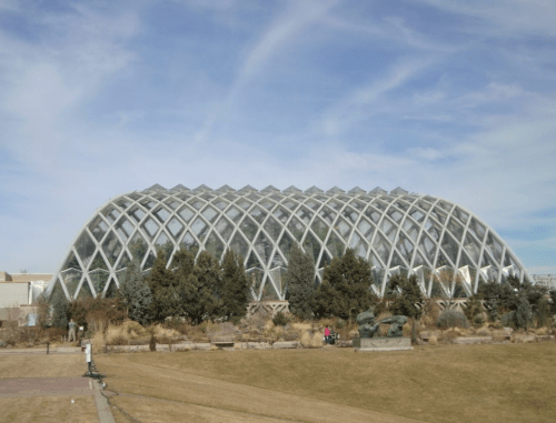 A large, modern glass greenhouse with a geometric design, surrounded by grass and trees under a blue sky.