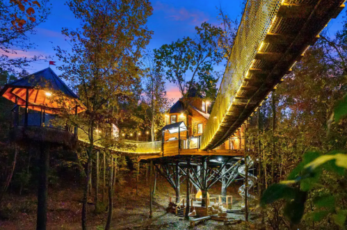 A treehouse with a bridge, illuminated at dusk, surrounded by trees and a colorful sky.