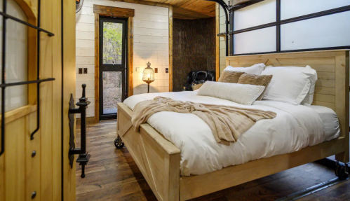 Cozy bedroom with a wooden bed, soft bedding, and warm lighting, featuring rustic decor and large windows.