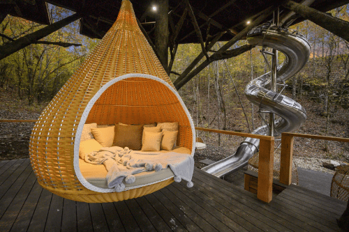 A cozy wicker hanging bed with cushions, next to a silver slide in a wooded outdoor setting.
