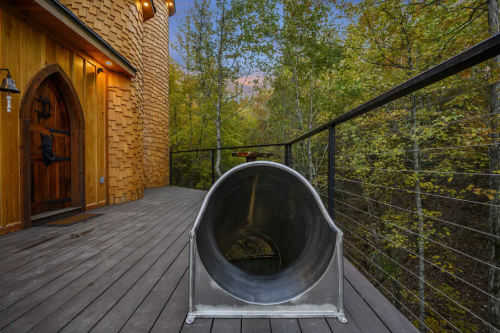 A metal slide on a wooden deck surrounded by trees, leading to a rustic wooden building.