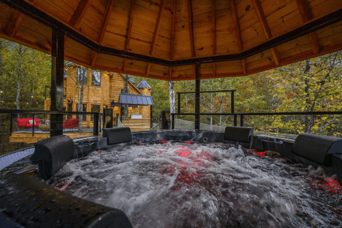 A hot tub with swirling water, surrounded by a wooden gazebo and trees, with a cabin visible in the background.