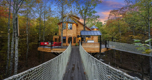 A wooden treehouse with a deck, surrounded by trees, connected by a rope bridge, under a colorful sunset sky.