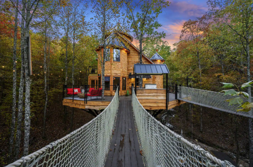 A wooden treehouse with a deck, surrounded by trees, connected by a rope bridge, under a colorful sunset sky.