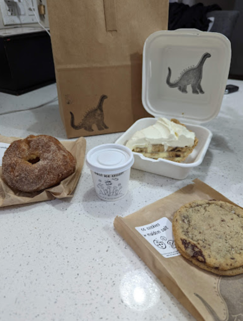 A bag and containers with a donut, cookie, and dessert topped with whipped cream on a kitchen counter.