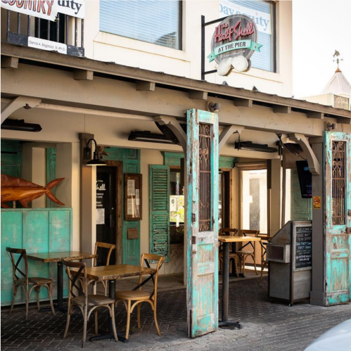 A rustic restaurant exterior with turquoise doors, wooden tables, and a fish decoration, set by the pier.