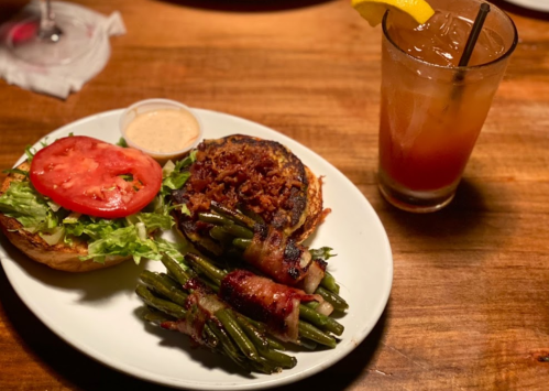 A plate with a burger topped with lettuce, tomato, and bacon-wrapped green beans, alongside a drink with a lemon slice.