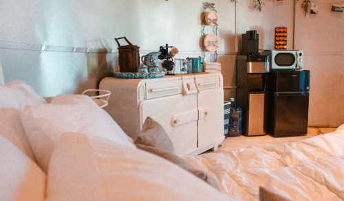 Cozy interior of a room featuring a bed, vintage dresser, microwave, and mini fridge against a softly lit wall.