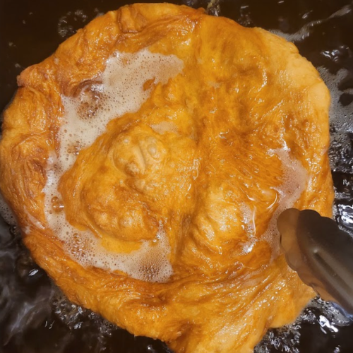 A golden-brown fried dough piece bubbling in hot oil, with tongs resting beside it.