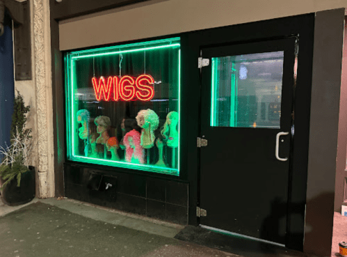 A storefront with a neon "WIGS" sign, showcasing colorful wigs in the window.