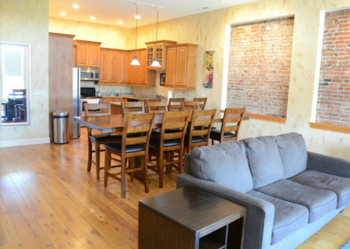Spacious kitchen and living area with wooden furniture, brick walls, and large windows, featuring a cozy gray sofa.