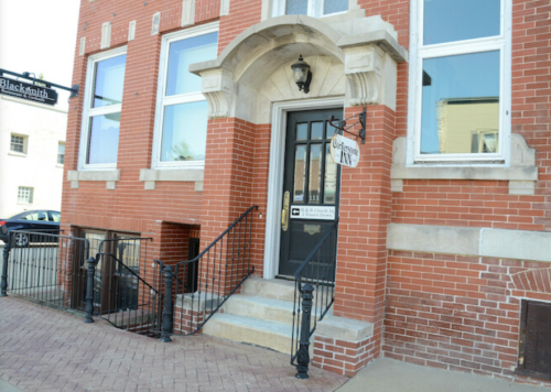 Brick building entrance with steps, a lantern, and a sign reading "Blacksmith." Sidewalk and railing visible.