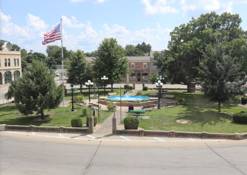 A park with a fountain, benches, and trees, featuring an American flag waving in the background under a blue sky.