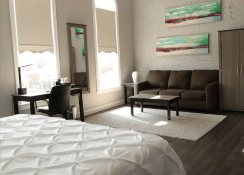 A cozy hotel room featuring a bed, sofa, coffee table, desk, and large windows with soft natural light.
