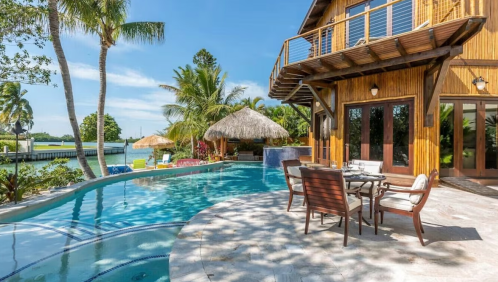 Tropical poolside view with a wooden house, palm trees, and a thatched gazebo under a clear blue sky.