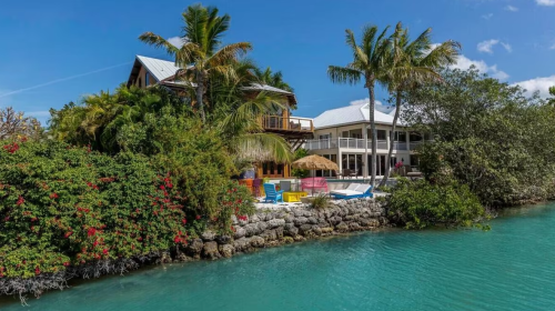 Tropical waterfront property with palm trees, colorful furniture, and a clear blue lagoon under a bright blue sky.