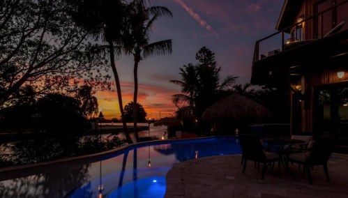 A serene sunset over a poolside with palm trees, reflecting vibrant colors in the sky and water.