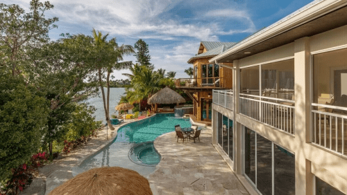 A luxurious backyard with a pool, palm trees, and a waterfront view, featuring a thatched-roof gazebo and modern home.