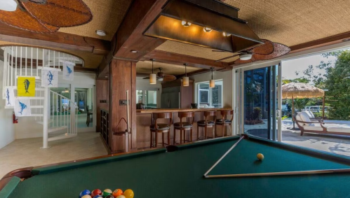 A spacious game room with a pool table, bar area, and large windows overlooking a patio and outdoor seating.