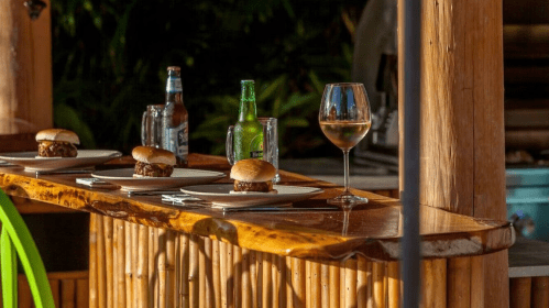 Three sliders on plates with drinks, including a beer and a glass of white wine, on a bamboo bar.