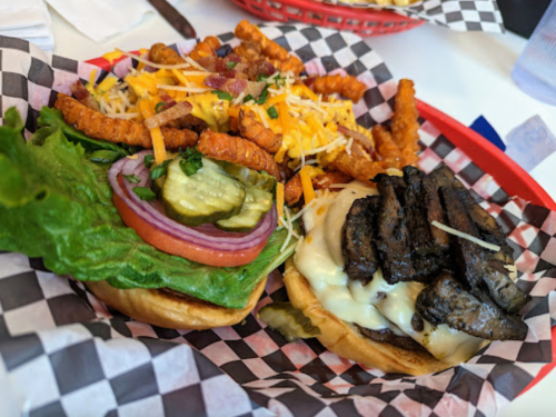 A close-up of a burger with lettuce, tomato, pickles, and cheese, served with a side of seasoned fries in a basket.