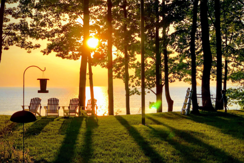 A serene lakeside scene at sunset, featuring chairs, trees, and a bird feeder casting long shadows on the grass.
