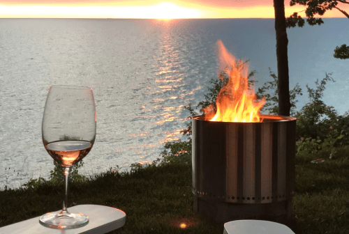 A glass of rosé wine beside a fire pit, with a sunset over a calm lake in the background.