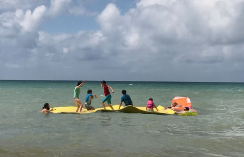 Children playing on a large float in the ocean, with a cloudy sky and gentle waves in the background.