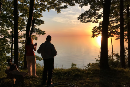 Silhouetted figures stand by a lake at sunset, surrounded by trees, capturing the serene view.