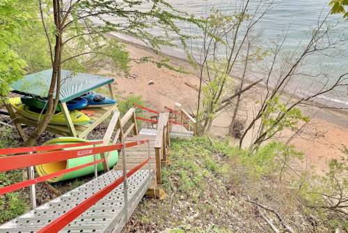 A wooden staircase leads down to a sandy beach, with kayaks stored under a shelter among green trees.