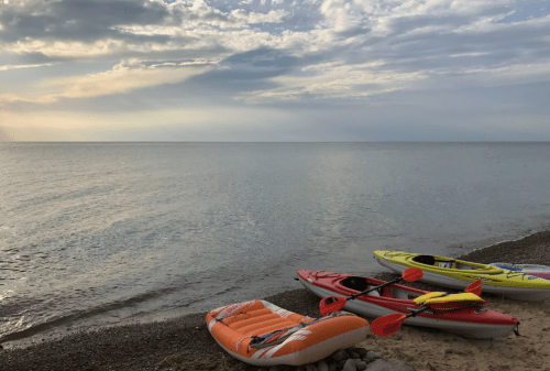 Colorful kayaks and a paddleboard on a sandy shore, with calm water and a cloudy sky in the background.