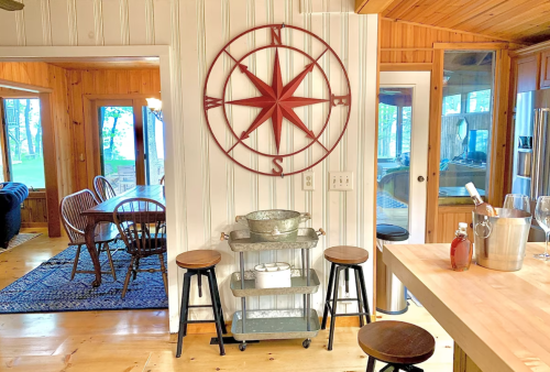 A cozy kitchen with a compass wall decor, bar cart, and wooden stools, leading to a dining area with large windows.