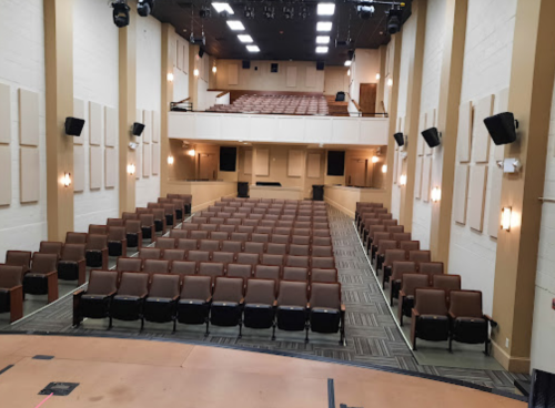 A spacious auditorium with rows of brown seats, stage in the foreground, and a balcony above.