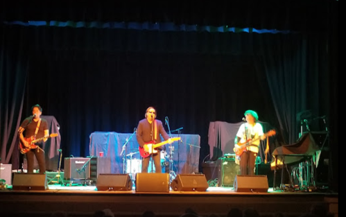 A band performs on stage with instruments, under colorful lights, in front of a dark backdrop.