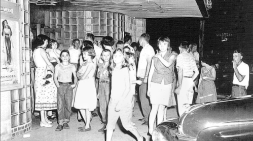 A crowd of people, mostly teenagers, gathered outside a theater at night, with vintage cars parked nearby.