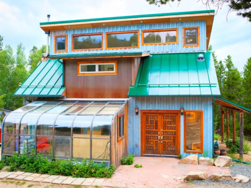 A charming two-story house with a green metal roof, large windows, and a greenhouse in front, surrounded by greenery.