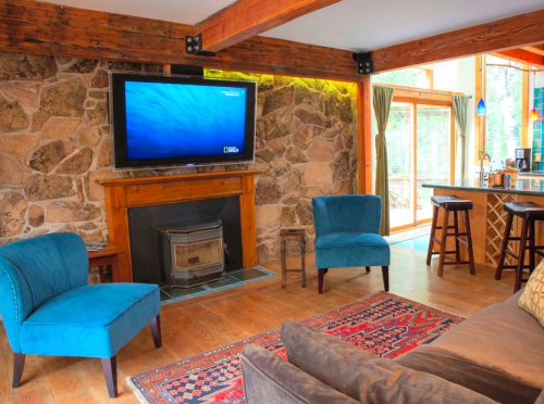 Cozy living room with stone walls, blue chairs, a TV above a fireplace, and a view of a kitchen area.