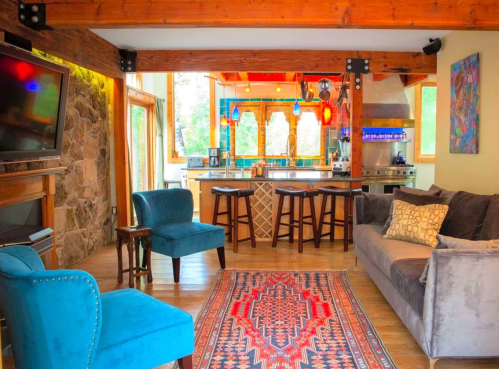 Cozy living area with a blue chair, gray sofa, colorful rug, and a kitchen in the background featuring wooden beams and bright lights.