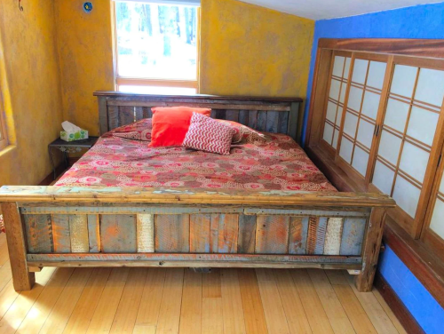 A rustic wooden bed with a colorful patterned blanket and pillows, set in a bright, cozy room with natural light.