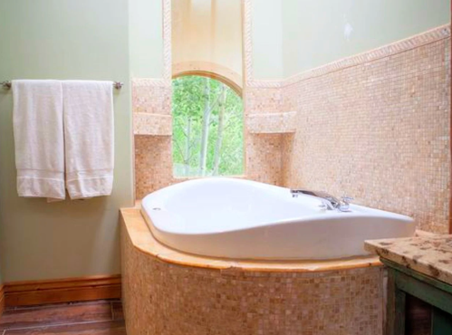 A modern bathroom featuring a curved white bathtub, beige mosaic tiles, and a window with a view of greenery.