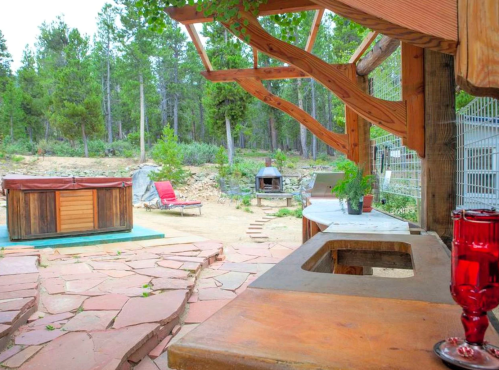 A rustic outdoor space with a wooden counter, hot tub, lounge chair, and lush greenery in the background.