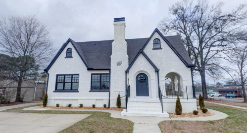 A charming white house with a dark roof, front steps, and landscaped yard, set against a cloudy sky.