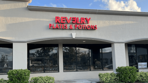 Sign for "Revelry Plates & Potions" on a building exterior, with large windows and greenery in front.
