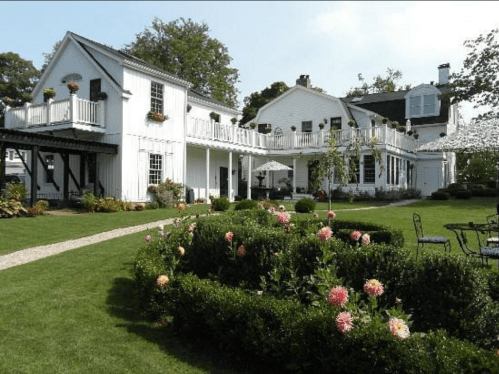 A charming white house with balconies, surrounded by lush gardens and colorful flowers on a sunny day.