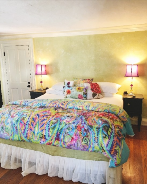 Brightly decorated bedroom with a colorful quilt, two lamps, and floral pillows against a light green wall.